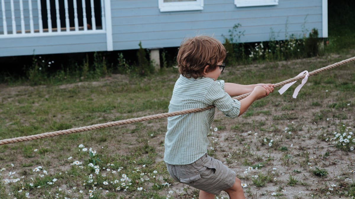 Young boy engaged in a tug of war outside near a blue building during the day.