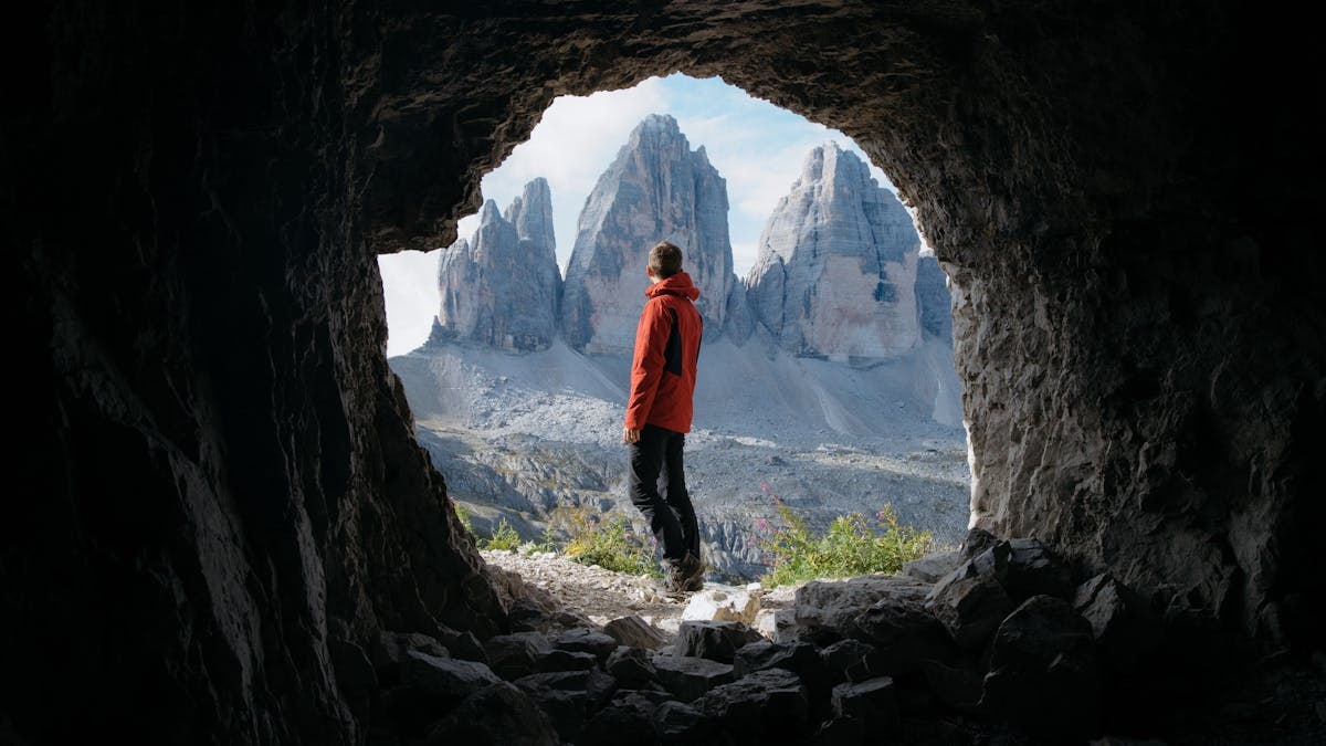 Man in red jacket standing in cave entrance, viewing Tre Cime di Lavaredo in Italy.